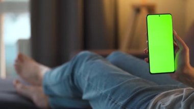 Evde bir kadın kanepeye uzanıyor ve dikey modda yeşil ekran ile akıllı telefon kullanıyor. İnternette gezinen, içerik izleyen, videolar, bloglar. POV.