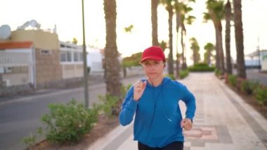 Kulaklıklı bir kadın gün batımında Palm Bulvarı boyunca koşuyor. Sağlıklı aktif yaşam tarzı. Yavaş çekim