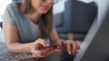 Kadın yerde yatıyor ve bir kredi kartı ve dizüstü bilgisayar kullanarak online alışveriş yapıyor. İnternetten alışveriş, yaşam tarzı teknolojisi.