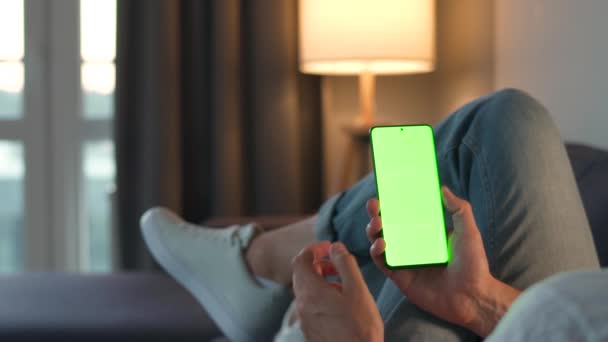 Mann zu Hause auf einem Sofa liegend und Smartphone mit grünem Bildschirm im vertikalen Modus. Er surft im Internet, schaut sich Inhalte, Videos, Blogs an. POV. — Stockvideo