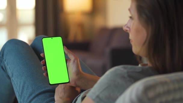 Vrouw thuis liggend op een bank en met behulp van smartphone met groene mock-up scherm in verticale modus. Meisje surfen op internet, kijken naar inhoud, video 's, blogs. — Stockvideo