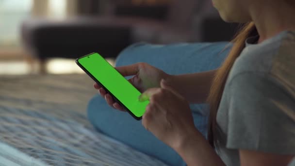 Женщина дома лежит на диване и использует смартфон с зеленым макетом экрана в вертикальном режиме. Девочки просматривают Интернет, смотрят контент, видео, блоги. — стоковое видео