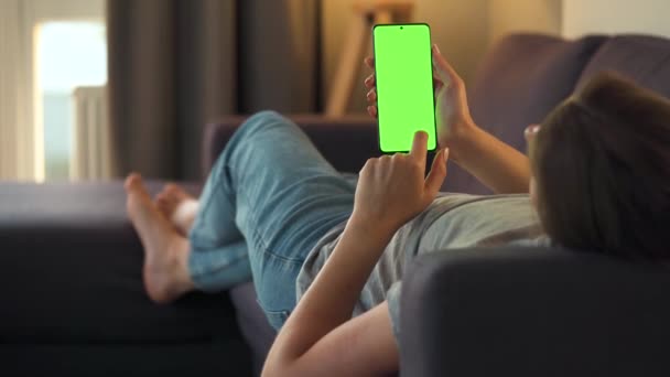Evde bir kadın kanepeye uzanıyor ve dikey modda yeşil ekran ile akıllı telefon kullanıyor. Kız internette geziniyor, içeriği, videoları, blogları izliyor. — Stok video