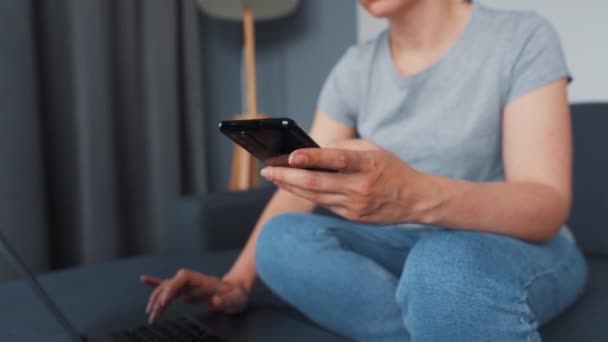 Frau sitzt auf einem gemütlichen Sofa und arbeitet an Laptop und Smartphone. Konzept der Fernarbeit. — Stockvideo