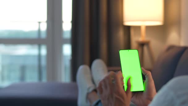 Man thuis liggend op een bank en met behulp van smartphone met groene mock-up scherm in verticale modus. Een man die op internet surft, inhoud, video 's, blogs bekijkt. POV. — Stockvideo