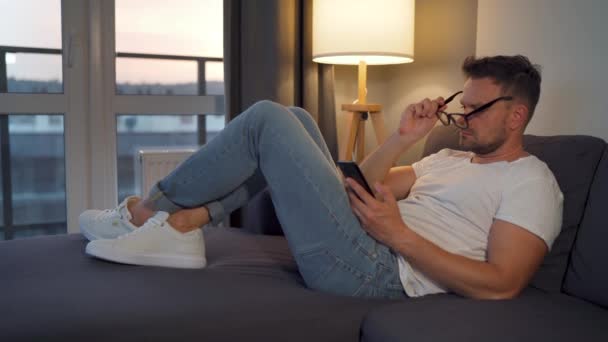 Homme avec des lunettes couché sur le canapé dans une chambre confortable et en utilisant un smartphone pour surfer sur Internet ou bavarder dans les messagers le soir. Concept de détente et de style de vie — Video