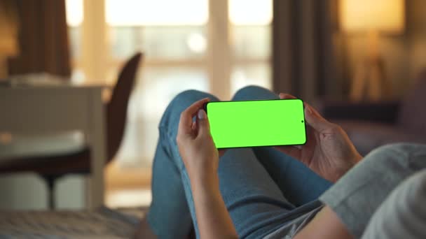 Женщина дома лежит на диване и использует смартфон с зеленым макетом экрана в горизонтальном режиме. Девушка просматривает Интернет, смотрит контент, видео, блоги. POV. — стоковое видео
