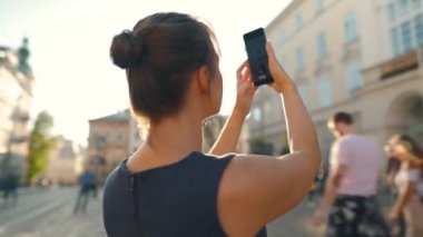 Kadın eski bir sokakta duruyor ve gün batımında akıllı bir telefonla fotoğraf ya da video çekiyor.