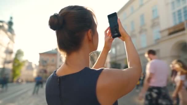 Mulher fica em uma rua velha e tira uma foto ou vídeo em um smartphone ao pôr do sol — Vídeo de Stock