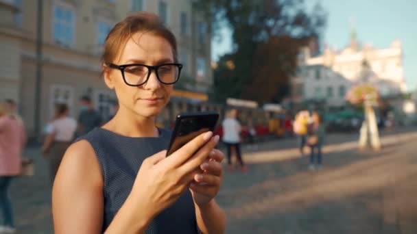 Eski bir sokakta duran, akıllı telefon kullanan ve etrafa bakan bir kadın. — Stok video