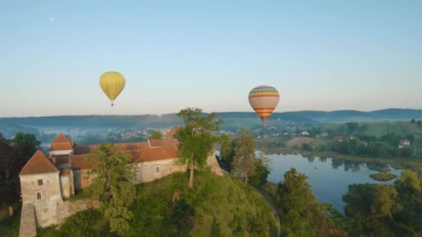 Barevné horkovzdušné balóny létají nad středověkým hradem a jezerem v ranní mlze. Manévrovatelný let a energetická smyčka. Cestování, dobrodružství, festival. — Stock video
