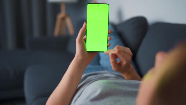 Žena doma leží na pohovce a používá smartphone se zelenou maketou obrazovky ve vertikálním režimu. Dívka prohlížení Internetu, sledování obsahu, videa, blogy. — Stock video