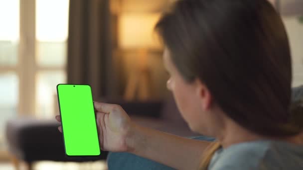Frau zu Hause auf dem Sofa liegend und Smartphone mit grünem Bildschirm im vertikalen Modus. Mädchen surfen im Internet, schauen Inhalte, Videos, Blogs.