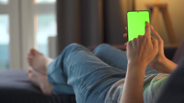 Vrouw thuis liggend op een bank en met behulp van smartphone met groene mock-up scherm in verticale modus. Meisje surfen op internet, kijken naar inhoud, video 's, blogs. POV. — Stockvideo