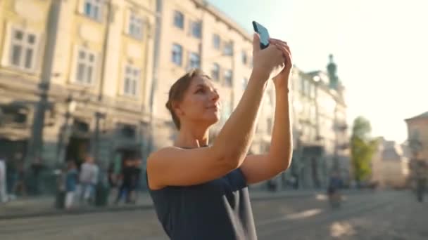 Mulher fica em uma rua velha e tira uma foto ou vídeo em um smartphone ao pôr do sol — Vídeo de Stock