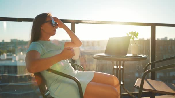 Mujer bebe café y descansa durante un descanso en el balcón contra el sol poniente. Concepto de trabajo remoto — Vídeos de Stock