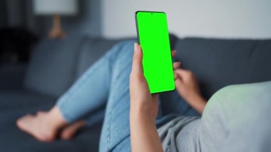 Evde bir kadın kanepeye uzanıyor ve dikey modda yeşil ekran ile akıllı telefon kullanıyor. İnternette gezinen, içerik izleyen, videolar, bloglar. POV.