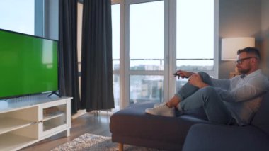 Evdeki adam kanepede uzanıp yeşil ekran TV izliyor.