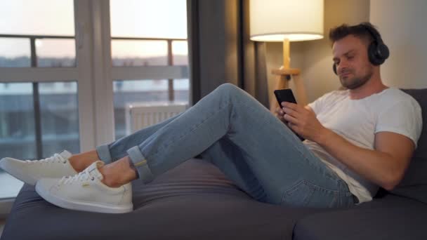 Ο άνθρωπος που βρίσκεται στον καναπέ σε ένα άνετο δωμάτιο, βάζει ακουστικά και χρησιμοποιώντας ένα smartphone για να ακούσετε μουσική. έννοια χαλάρωσης και τρόπου ζωής — Αρχείο Βίντεο