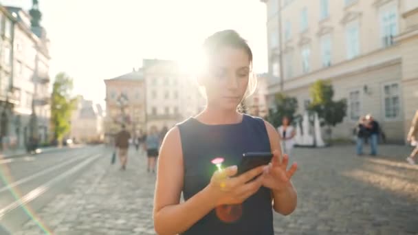 Eski bir caddede yürüyen ve günbatımında akıllı telefon kullanan bir kadın. — Stok video