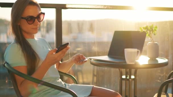 Nő ül az erkélyen hátterében a lenyugvó nap, és egy online vásárlás hitelkártyával és okostelefonnal — Stock videók
