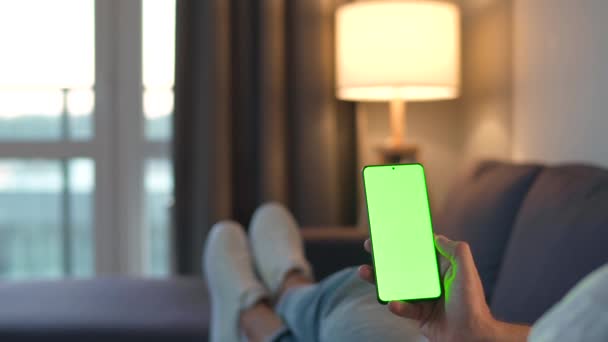 自宅の男はソファに横になり、垂直モードで緑色のモックアップ画面を持つスマートフォンを使用しています。男は、コンテンツ、ビデオ、ブログを見て、インターネットを閲覧。POV. — ストック動画