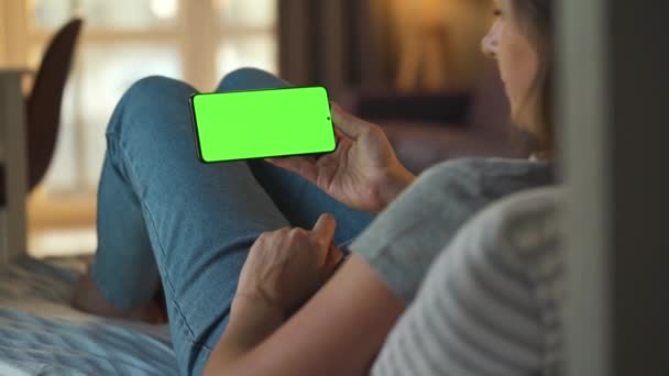 Vrouw thuis liggend op een bank en met behulp van smartphone met groene mock-up scherm in horizontale modus. Meisje surfen op internet, kijken naar inhoud, video 's, blogs. — Stockvideo