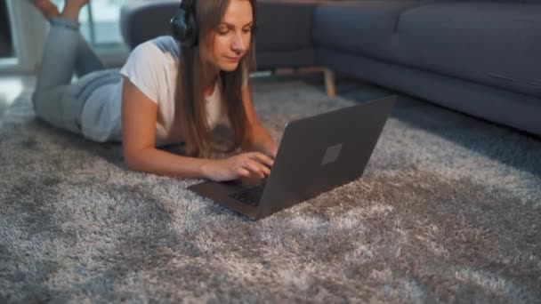 Frau liegt auf dem Boden, arbeitet an einem Laptop und hört Musik über Kopfhörer. Konzept der Fernarbeit. — Stockvideo