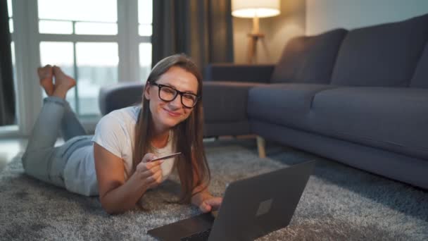 Mujer está tirado en el suelo y y hace una compra en línea con una tarjeta de crédito y un ordenador portátil. Compras en línea, tecnología de estilo de vida — Vídeos de Stock