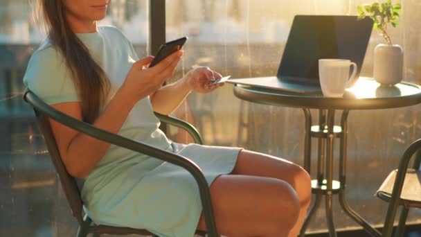 Vrouw zittend op het balkon tegen de achtergrond van de ondergaande zon en doet een online aankoop met een creditcard en smartphone — Stockvideo