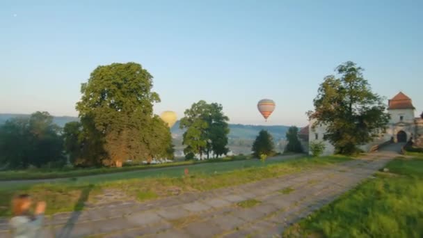 Kolorowe balony na ogrzane powietrze przelatują nad średniowiecznym zamkiem i jeziorem w porannej mgle. Kobieta robi zdjęcia na zamku smartfona i balony — Wideo stockowe