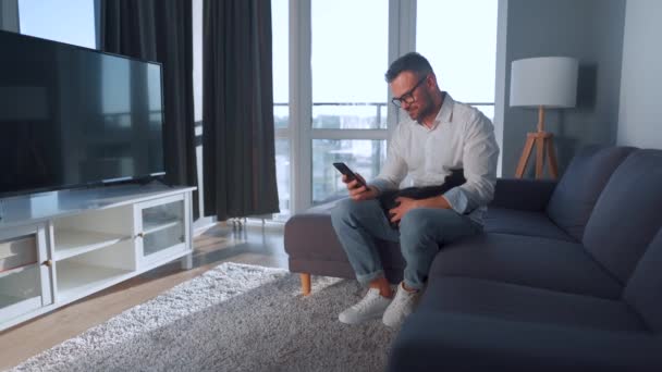 Man met bril zittend op de bank in een gezellige kamer en met smartphone surfen op internet of chatten in boodschappers. Hij streelt een zwarte kat die op zijn schoot ligt.. — Stockvideo