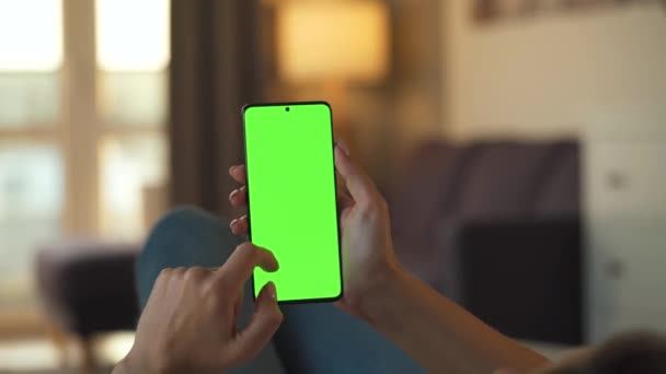 Mulher em casa deitada em um sofá e usando smartphone com tela verde mock-up no modo vertical. Menina navegando na Internet, assistindo conteúdo, vídeos, blogs. VPO. — Vídeo de Stock