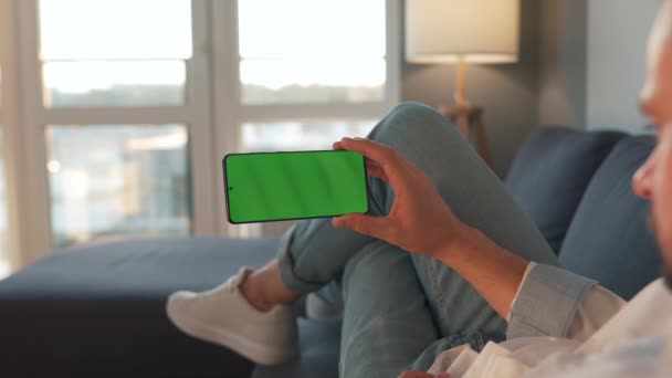 Человек дома лежит на диване и использует смартфон с зеленым макетом экрана в горизонтальном режиме. Он просматривает Интернет, смотрит контент, видео, блоги — стоковое видео