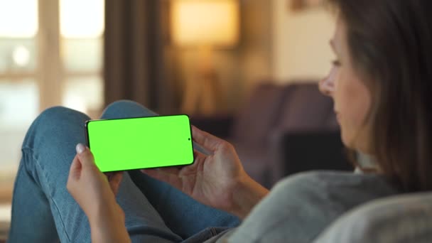 Frau zu Hause auf dem Sofa liegend und Smartphone mit grünem Bildschirm im horizontalen Modus. Mädchen surfen im Internet, schauen Inhalte, Videos, Blogs. — Stockvideo