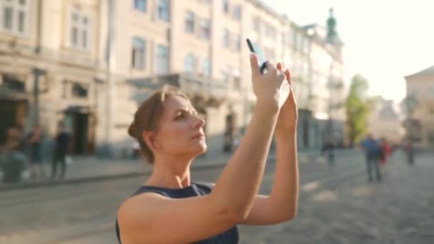 Mulher fica em uma rua velha e tira uma foto ou vídeo em um smartphone ao pôr do sol — Vídeo de Stock