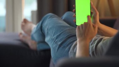 Evde bir kadın kanepeye uzanıyor ve dikey modda yeşil ekran ile akıllı telefon kullanıyor. İnternette gezinen, içerik izleyen, videolar, bloglar. POV.