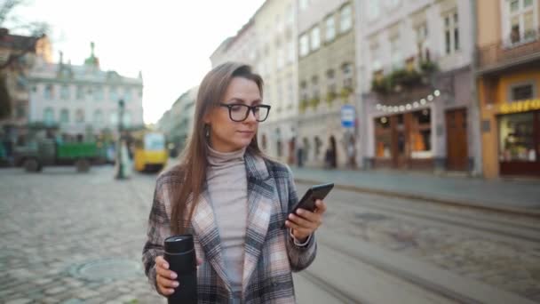 Mulher de casaco e óculos com uma xícara térmica nas mãos fala em um smartphone enquanto caminha na praça da cidade. O eléctrico amarelo está a passar. Arquitetura europeia velha em torno. — Vídeo de Stock