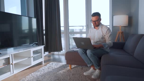 Man met bril zittend op de bank in een gezellige kamer en met laptop. Afstandswerkconcept — Stockvideo