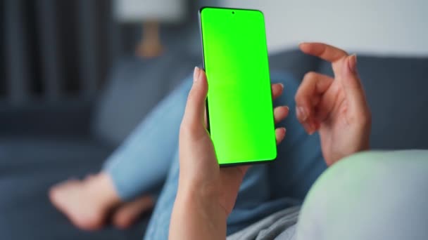 Mulher em casa deitada em um sofá e usando smartphone com tela verde mock-up no modo vertical. Menina navegando na Internet, assistindo conteúdo, vídeos, blogs. VPO. — Vídeo de Stock
