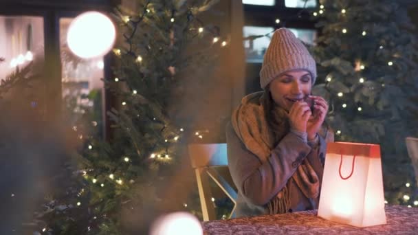 La mujer se sienta en una terraza al aire libre en invierno y mira en una bolsa con un regalo y se regocija por lo que encuentra allí. El paquete se destaca desde el interior — Vídeos de Stock