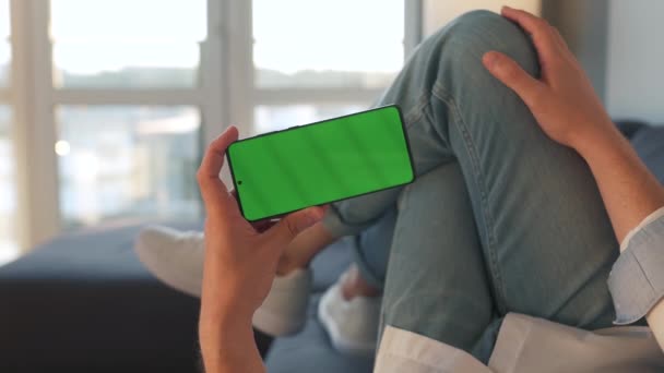 Man hemma liggande på en soffa och använda smartphone med grön mock-up skärm i horisontellt läge. Han surfar på Internet, tittar på innehåll, videor, bloggar — Stockvideo