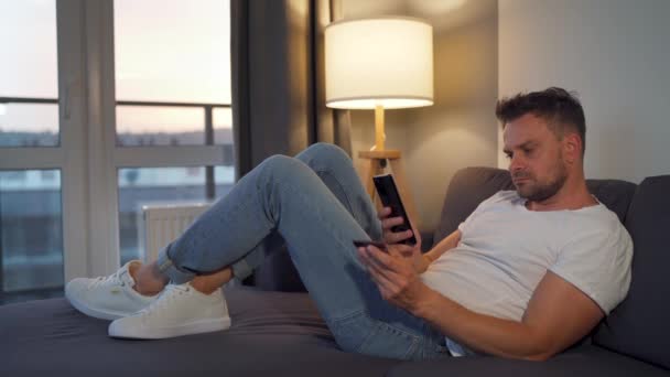 Mężczyzna leżący na kanapie w przytulnym pokoju i dokonujący zakupów online za pomocą karty kredytowej i jej smartfona. Zakupy online, technologia stylu życia. — Wideo stockowe
