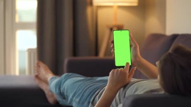 Evde bir kadın kanepeye uzanıyor ve dikey modda yeşil ekran ile akıllı telefon kullanıyor. Kız internette geziniyor, içeriği, videoları, blogları izliyor.
