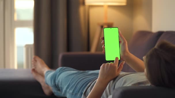 Γυναίκα στο σπίτι ξαπλωμένη σε καναπέ και χρησιμοποιώντας smartphone με πράσινη οθόνη mock-up σε κάθετη λειτουργία. Κορίτσι περιήγηση στο Internet, παρακολουθώντας περιεχόμενο, βίντεο, blogs. — Αρχείο Βίντεο