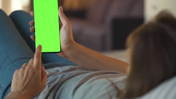 Frau zu Hause auf dem Sofa liegend und Smartphone mit grünem Bildschirm im vertikalen Modus. Mädchen surfen im Internet, schauen Inhalte, Videos, Blogs. — Stockvideo