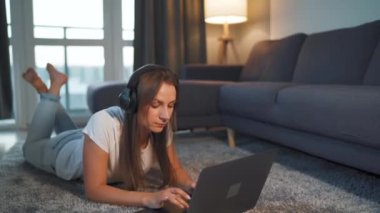 Kadın yerde yatıyor, dizüstü bilgisayarla çalışıyor ve kulaklıkla müzik dinliyor. Uzak çalışma kavramı.