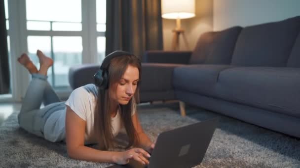 La femme est allongée sur le sol, travaille sur un ordinateur portable et écoute de la musique sur un casque. Concept de télétravail. — Video