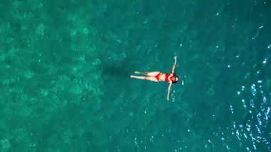Kırmızı mayo giymiş bir kadının havadan görünüşü, kristal berrak deniz sularında rahatlama somutlaştırıcı bir şekilde zahmetsizce sırt üstü yüzüyor. Yaz tatilleri.