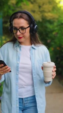 Gözlüklü ve kulaklıklı mutlu kadın elinde kahve ve akıllı telefonuyla çiçek açan bahçede ya da yaz günü yeşil sokakta yürüyor..